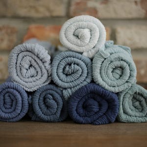 Puede incluir: Una pila de toallitas enrolladas en varios tonos de azul y gris. Las toallitas están dispuestas en forma de pirámide, con el azul más oscuro en la base y tonos más claros hacia arriba. Se aprecia la textura del tejido.