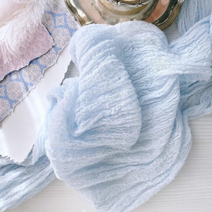 May include: A pale blue, textured fabric is arranged on a white surface, with a silver tray holding dried flowers in the background. A white feather and patterned paper are also present, creating a soft, tactile aesthetic.