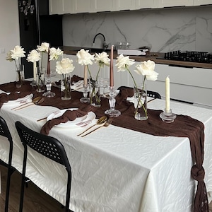 Könnte beinhalten: Ein Esstisch für einen besonderen Anlass. Der Tisch ist mit einer weißen Tischdecke und einem braunen Tischläufer bedeckt. Weiße Rosen in Glasvasen, Kerzen und goldfarbenes Besteck vervollständigen die elegante Einrichtung. Schwarze Stühle sind sichtbar.