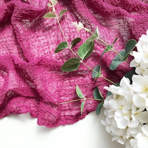 Puede incluir: Primer plano de una tela magenta texturizada y arrugada. Ramas verdes y un racimo de flores blancas están dispuestas sobre la tela, creando una presentación decorativa. La tela se encuentra sobre un fondo blanco.