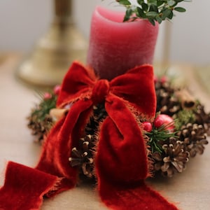 May include: A dark pink pillar candle sits in the center of a circular wreath made of brown pinecones, green sprigs, and red berries. A large, deep red velvet ribbon with frayed edges is tied around the base of the candle and the wreath.