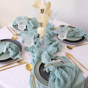 Könnte beinhalten: Elegante Tischdekoration mit türkisfarbenen Servietten, goldenem Besteck und klaren Kristallgläsern. Die Teller sind schwarz und hellgrün. Eine weiße Kerze und ein Blumengesteck dienen als Mittelstück.