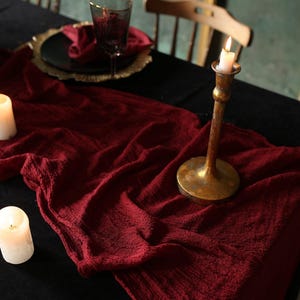 Puede incluir: Una mesa decorada con un mantel negro, un camino de mesa rojo oscuro texturizado y un candelabro de latón con una vela encendida. También hay una copa de vino, un plato y una servilleta. La escena evoca un ambiente cálido y acogedor.