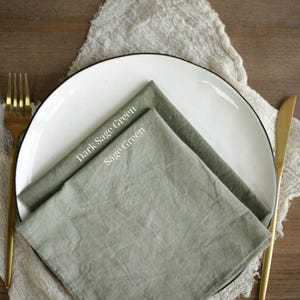 May include: A white plate with a black rim displays two sage green napkins, labelled "Dark Sage Green" and "Sage Green." A gold fork and knife flank the plate, set on a textured, off-white cloth.
