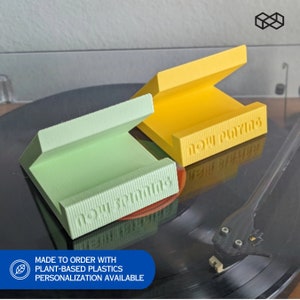 May include: Two 3D printed record risers in green and yellow. The risers are angled and have the text "now playing" printed on them. The risers are on a black turntable with a needle.