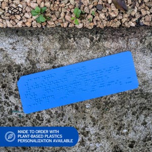 May include: A bright blue rectangular object with raised dots, likely a Braille product. The object is made of plant-based plastics. The text on the object is not clear. The background is a rough concrete surface with small rocks and greenery.
