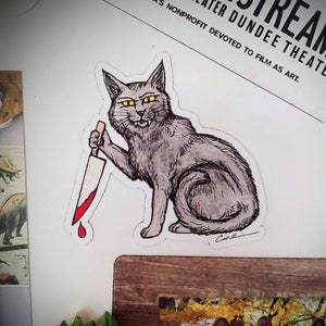May include: A sticker of a gray cat holding a bloody knife. The cat has yellow eyes and a menacing expression. The sticker is on a white background.
