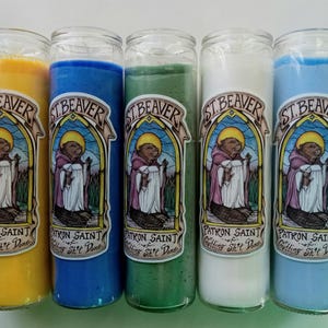 May include: Five glass jar candles in yellow, blue, green, white, and light blue. Each candle has a label with the text "ST. BEAVER PATRON SAINT Getting Shit Done" and an illustration of a beaver in a robe.