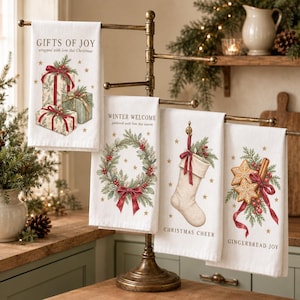 May include: Four white kitchen towels hanging on a bronze metal stand. Each towel features festive illustrations: stacked gifts with "GIFTS OF JOY", a wreath with "WINTER WELCOME", a stocking with "CHRISTMAS CHEER", and gingerbread with "GINGERBREAD JOY".