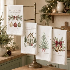 May include: Four white kitchen towels hanging on a bronze metal stand. Each towel features a unique Christmas-themed illustration: hanging ornaments with the text PEACE & JOY, a house with the text HOME FOR CHRISTMAS, a pine tree with MERRY & BRIGHT, and bells with CHRISTMAS MAGIC.