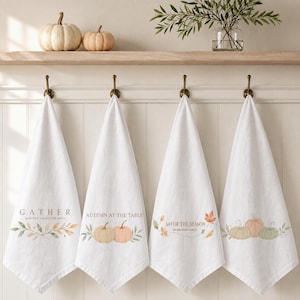 May include: Four white kitchen towels hanging on brass hooks. Each towel features watercolor illustrations of pumpkins and autumn leaves. The text reads GATHER, AUTUMN AT THE TABLE, SAVOR THE SEASON, and includes small phrases under the main titles.