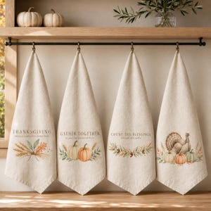 May include: Four beige kitchen towels hanging from a black metal rod. Each towel features autumn-themed illustrations and text: THANKSGIVING, GATHER TOGETHER, COUNT THE BLESSINGS, and a turkey with pumpkins. The designs include wheat, leaves, and acorns.