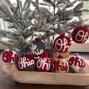 May include: A selection of Ohio-themed Christmas baubles in a wooden bowl. The baubles are red, grey, and green, with the word "Ohio" in white script. Some baubles are hanging from a frosted Christmas tree.