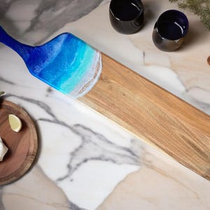May include: Wooden serving board with a blue resin ocean design. The handle is solid blue, transitioning to a beach scene with white and turquoise waves. The board is approximately 24 inches long.