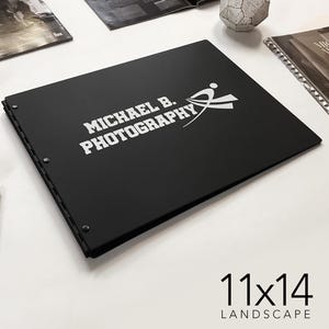 以下が含まれることがあります： 黒い写真アルバム。白いロゴには「MICHAEL B. PHOTOGRAPHY」とあり、白いシルエットでボートを漕ぐ人が描かれています。アルバムのサイズは11x14インチで、横向きです。