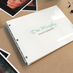 May include: A white photo album with a clear plastic cover. The album has a silver ring binder and the text "Dee Murphy PHOTOGRAPHY" is printed on the front cover.