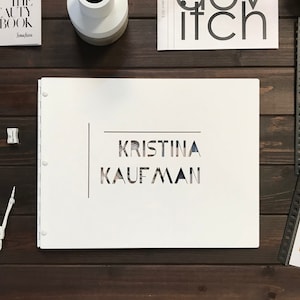Puede incluir: Una carpeta blanca de tres anillos con el nombre "KRISTINA KAUFMAN" recortado en un patr&oacute;n geom&eacute;trico.