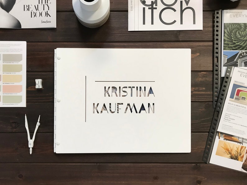 Puede incluir: Una carpeta blanca de tres anillos con el nombre "KRISTINA KAUFMAN" recortado en un patr&oacute;n geom&eacute;trico.