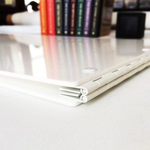 Puede incluir: Carpeta de pl&aacute;stico blanco con una bisagra de metal. La carpeta est&aacute; cerrada y se encuentra sobre una superficie blanca.