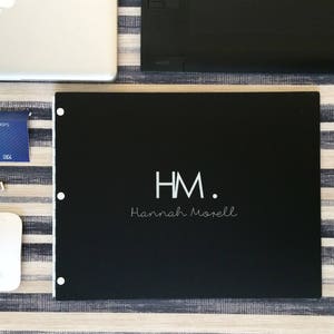 K&ouml;nnte beinhalten: Ein schwarzes Portfolio mit den Initialen "HM" und dem Namen "Hannah Morell" in wei&szlig;. Das Portfolio liegt auf einer gestreiften Oberfl&auml;che.