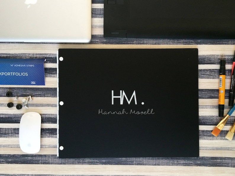 K&ouml;nnte beinhalten: Ein schwarzes Portfolio mit den Initialen "HM" und dem Namen "Hannah Morell" in wei&szlig;. Das Portfolio liegt auf einer gestreiften Oberfl&auml;che.