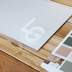 May include: A white acrylic sign with the letters "LG" embossed in white. The sign is on a wooden surface.