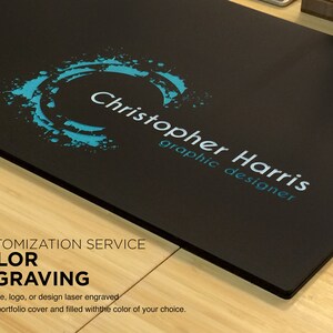 K&ouml;nnte beinhalten: Ein schwarzer Portfolio-Umschlag mit einem blauen und wei&szlig;en Logo, auf dem "Christopher Harris graphic designer" steht. Der Umschlag bietet einen "Farbgravur"-Service, bei dem Sie Ihren Namen, Ihr Logo oder Ihr Design lasergravieren und mit der Farbe Ihrer Wahl f&uuml;llen lassen k&ouml;nnen.