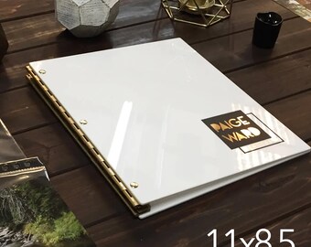 Puede incluir: Álbum de fotos blanco con bisagras doradas y detalles en latón. El álbum está cerrado y tiene una pegatina de lámina dorada con el texto "Paige Ward" en la parte delantera. El álbum está sobre una superficie de madera. 11x8.5 retrato.