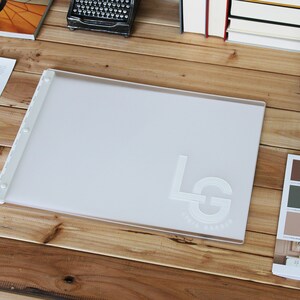 May include: A white plastic presentation board with a raised, embossed logo that says "LG" in a circle. The board is on a wooden table with other items, including a book, a typewriter, a cup of coffee, and a paint color swatch book.