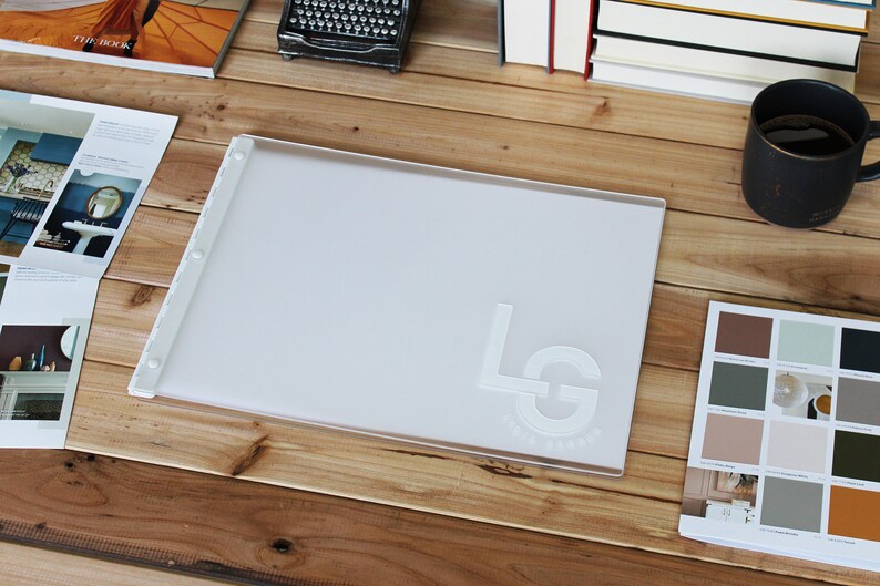 May include: A white plastic presentation board with a raised, embossed logo that says "LG" in a circle. The board is on a wooden table with other items, including a book, a typewriter, a cup of coffee, and a paint color swatch book.