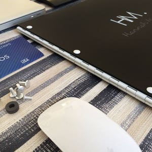 K&ouml;nnte beinhalten: Ein schwarzes, rechteckiges Portfolio mit einem silbernen Scharnier und wei&szlig;en Punkten. Das Portfolio tr&auml;gt die Initialen "H.M." und den Namen "Hannah Wadell" in wei&szlig; auf der Vorderseite.