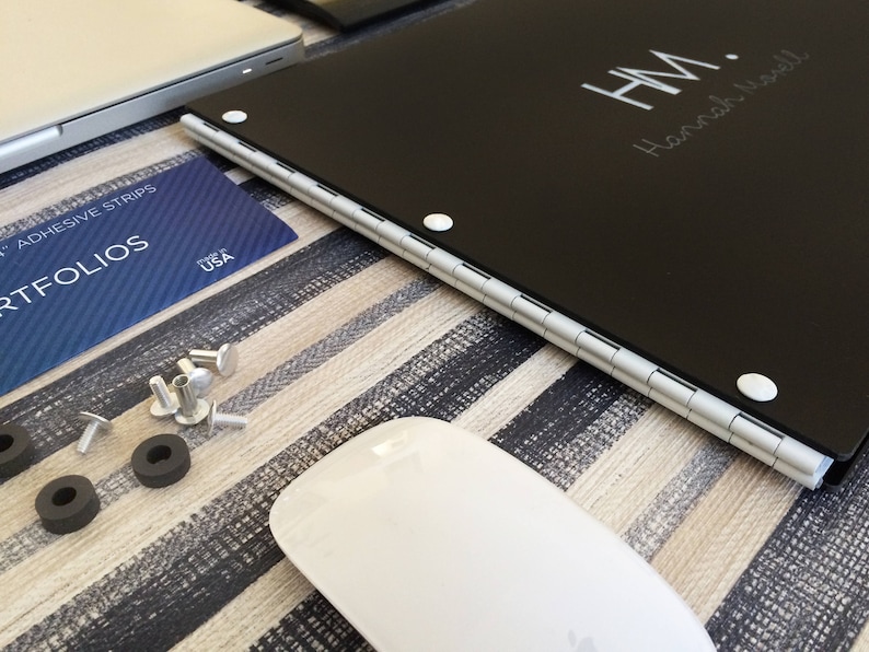 K&ouml;nnte beinhalten: Ein schwarzes, rechteckiges Portfolio mit einem silbernen Scharnier und wei&szlig;en Punkten. Das Portfolio tr&auml;gt die Initialen "H.M." und den Namen "Hannah Wadell" in wei&szlig; auf der Vorderseite.