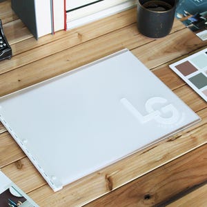 May include: A white portfolio with a white embossed "LG" logo on the front. The portfolio is lying open on a wooden table with other items, including a vintage typewriter, books, and a small tray of paint samples.