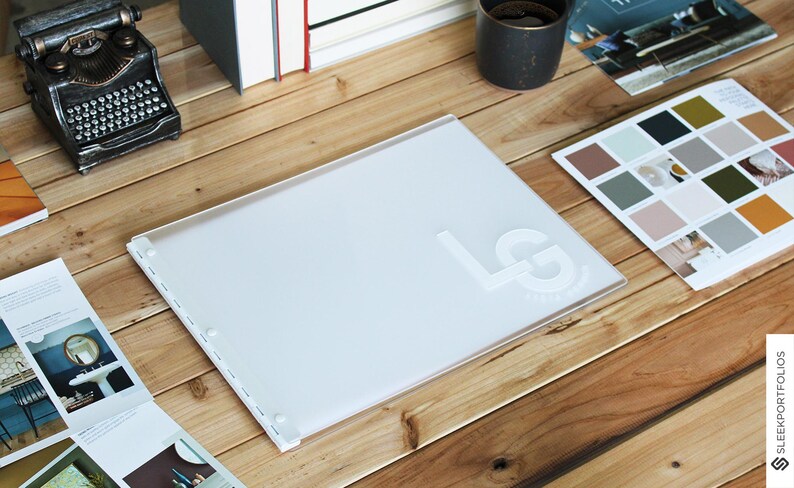 May include: A white portfolio with a white embossed "LG" logo on the front. The portfolio is lying open on a wooden table with other items, including a vintage typewriter, books, and a small tray of paint samples.