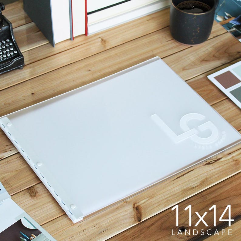 May include: A white plastic 11x14 inch landscape photo album with a clear cover and a white embossed "LG" logo. The album has a ring binding system and is lying on a wooden surface.