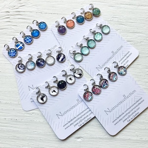 Peut inclure: Une collection de petits pendentifs ronds en métal présentant divers motifs colorés, incluant des formes géométriques, des motifs floraux et de l'art abstrait. Chaque pendentif possède une petite boucle argentée sur des cartes blanches.