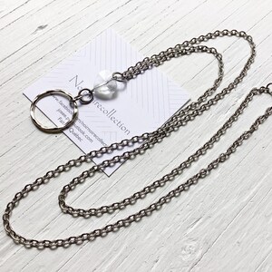 May include: A silver-toned metal chain necklace featuring a circular pendant and a clear, faceted crystal bead. The necklace is displayed on a white card with the text Noir collection, a Facebook URL, and the words Fait au Québec printed in black.