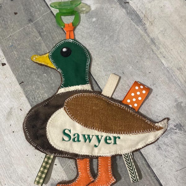 May include: A duck-shaped baby toy with a green soother attached. The duck features a green head, brown body, and orange feet. The name "Sawyer" is embroidered on the duck's chest. Various ribbons are attached to the duck.