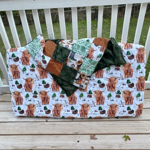 May include: A white crib sheet with a pattern of brown Highland cows and green trees. A green, brown, and white patchwork quilt is on top of the sheet.