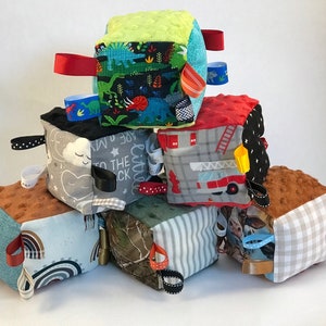 May include: A set of seven colorful fabric baby blocks with different patterns and textures. Each block has a ribbon loop for easy grasping. The blocks feature themes like dinosaurs, fire trucks, rainbows, and clouds.