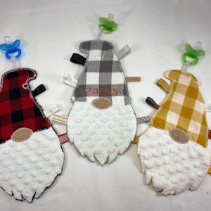 May include: Three handmade baby teethers shaped like gnomes. Each gnome has a different plaid fabric pattern: red and black, gray and white, and yellow and white. The gnomes have white fuzzy beards and a pacifier attached to the top.