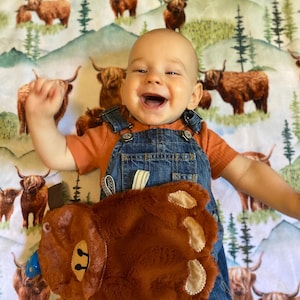May include: A smiling baby wearing denim overalls and an orange shirt, lying on a blanket with a highland cow pattern. The baby is holding a brown plush bear toy with cream-colored paw pads and a blue nose.