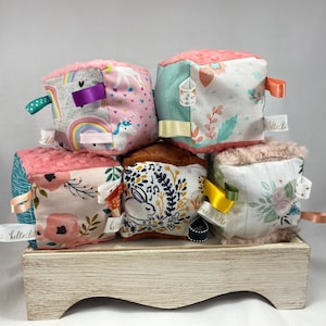 May include: A stack of six soft fabric baby blocks. Each block features a different patterned fabric, including florals, unicorns, and rainbows. The blocks have plush edges and colorful ribbons attached. Each block has a small tag that says "hello little".