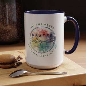 May include: White ceramic mug with a navy blue handle and interior rim. The mug features the text "PRATER Calligraphy" in a circular design with colorful accents. A spoon and a macaron are on a wooden surface.