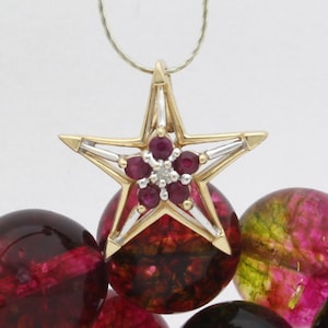 May include: A gold star-shaped pendant with a cluster of red rubies and a single diamond in the center. The pendant is hanging from a thin silver chain.