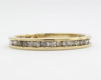 May include: A gold band ring with a row of small, round, gray gemstones set across the top. The ring is set against a plain white background, highlighting the ring's design and details.