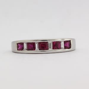May include: A silver ring featuring five square-cut, vibrant red gemstones, alternating with smaller, rectangular clear stones. The ring has a smooth, polished band, showcasing a classic and elegant design. The gemstones are set flush within the band.