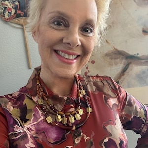 Puede incluir: Una mujer con una blusa roja con estampado floral y un collar en capas con colgantes cuadrados dorados. El collar presenta cuentas marrones y un cordón marrón oscuro. La blusa tiene cuello abotonado y mangas largas.