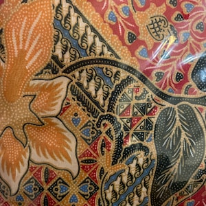 May include: A close-up of a fabric with a colorful batik pattern. The pattern features a mix of red, blue, black, and gold colors. The fabric has a traditional Indonesian design.