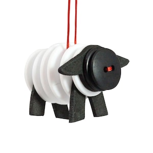 Può includere: Un ornamento a forma di pecora stravagante con un corpo bianco fatto di pezzi circolari impilati. La testa è un grande bottone nero con un centro rosso e le orecchie sono in feltro nero. La pecora poggia su zampe di feltro nero e pende da un filo rosso.